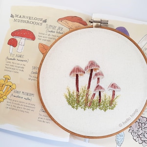 Könnte beinhalten: Gestickte Pilz-Darstellung auf einem weißen Stoff, der über einen hölzernen Stickrahmen gespannt ist. Der Rahmen hat einen Durchmesser von 25 cm. Die Pilze sind rosa und braun mit grünem Laub.