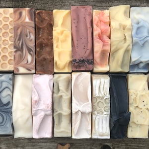 May include: A collection of 20 handmade soap bars in various colours and patterns, including swirls, honeycomb, and speckles. The bars are arranged in two rows of ten on a wooden surface.