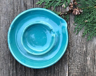Może przedstawiać: Turkusowa ceramiczna popielniczka o okrągłym kształcie. Popielniczka ma podniesiony środek i zakrzywioną krawędź do oparcia papierosów. Element jest szkliwiony błyszczącym wykończeniem i leży na drewnianej powierzchni.