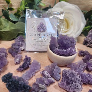 May include: A collection of purple grape agate crystal clusters scattered on a wooden surface, with some pieces inside a small round wooden bowl. A clear plastic bag contains a label with the text GRAPE AGATE FROM WEST SULAWESI INDONESIA.