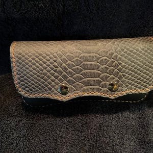 May include: A gray, textured wallet with a snakeskin pattern. The wallet has a rectangular shape with rounded edges and is secured with two silver-colored snaps. The edges are stitched with orange thread.