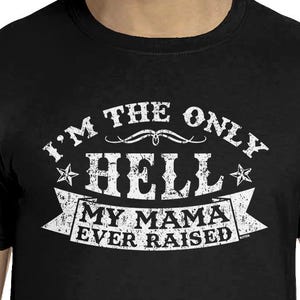 May include: A black short-sleeved t-shirt featuring white distressed-style text that reads I'M THE ONLY HELL MY MAMA EVER RAISED, centered on the chest. The design includes a banner graphic and two small stars flanking the word HELL.