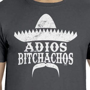 Op de afbeelding: Donkergrijs t-shirt met een witte sombrero-afbeelding, snor en de tekst "ADIOS BITCHACHOS". De afbeelding heeft een vintage uitstraling. Het t-shirt heeft korte mouwen en is gemaakt van zacht materiaal.