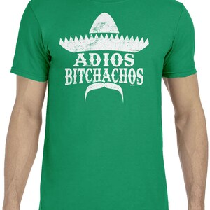 Op de afbeelding: Groen t-shirt met een witte sombrero-afbeelding, snor en de tekst "ADIOS BITCHACHOS". Het ontwerp is verweerd, waardoor het een vintage uitstraling heeft. Het t-shirt is effen groen van kleur.