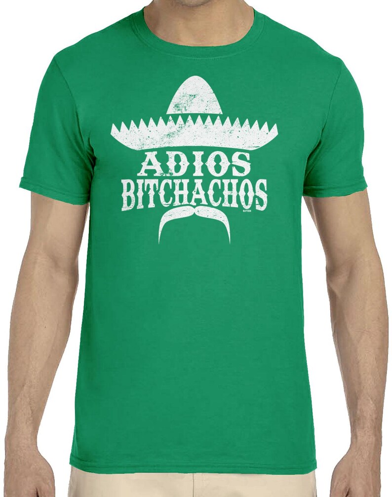 Op de afbeelding: Groen t-shirt met een witte sombrero-afbeelding, snor en de tekst "ADIOS BITCHACHOS". Het ontwerp is verweerd, waardoor het een vintage uitstraling heeft. Het t-shirt is effen groen van kleur.