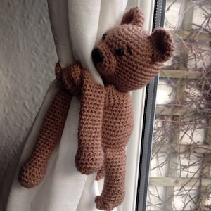 May include: A brown crocheted teddy bear with long arms is used as a curtain tieback. The bear is holding onto a white curtain with its arms.
