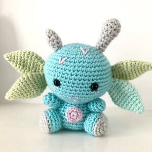 May include: A crocheted blue and green plush toy with large ears and a pink spot on its belly. The toy has black eyes and a stitched smile.