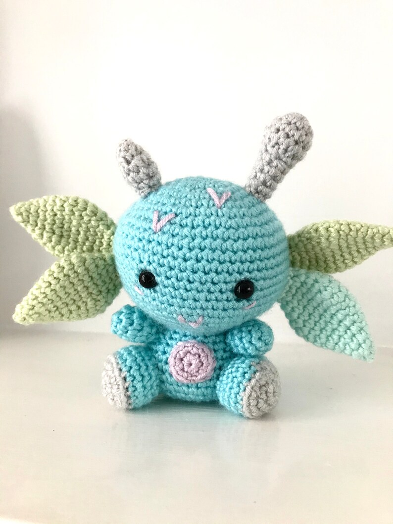 May include: A crocheted blue and green plush toy with large ears and a pink spot on its belly. The toy has black eyes and a stitched smile.