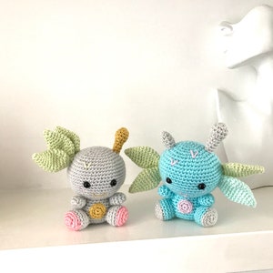 May include: Two crocheted plush toys with green wings and pink and yellow accents. The toys are sitting on a white shelf.