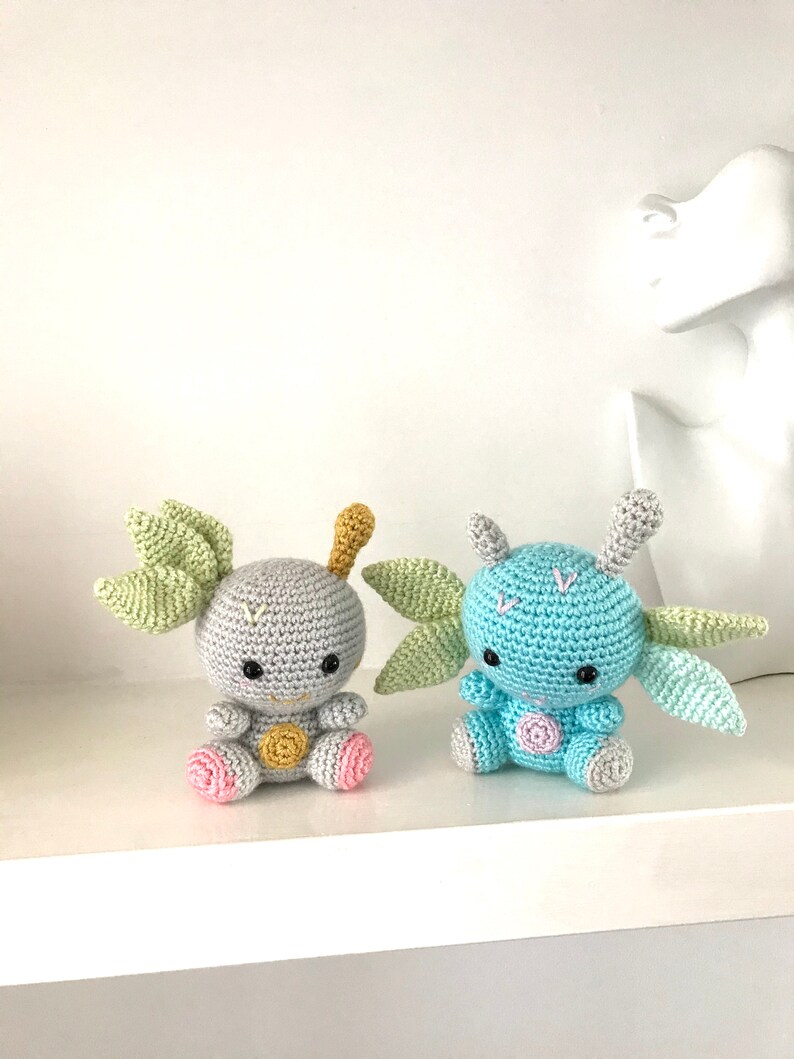 May include: Two crocheted plush toys with green wings and pink and yellow accents. The toys are sitting on a white shelf.