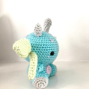 May include: A crocheted blue and green dinosaur toy with grey horns and feet. The dinosaur has a black eye and pink accents.
