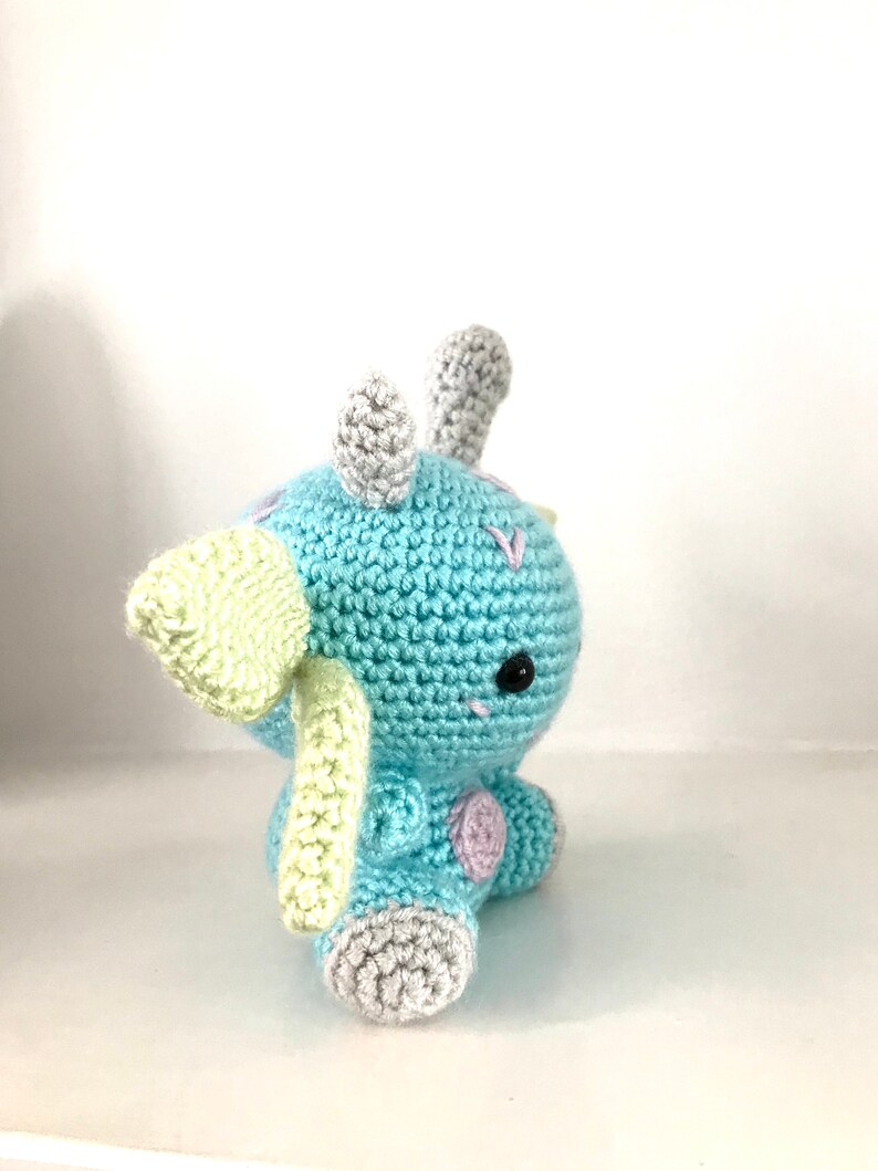 May include: A crocheted blue and green dinosaur toy with grey horns and feet. The dinosaur has a black eye and pink accents.