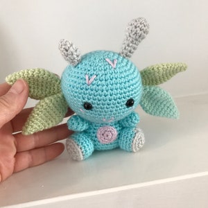 May include: A crocheted blue and green dragon toy with gray horns and a pink belly. The dragon is sitting on a white surface.