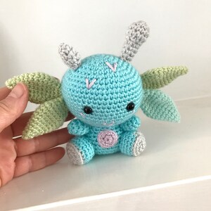 May include: A crocheted blue dragon with green wings and gray horns. The dragon has pink accents and black eyes.