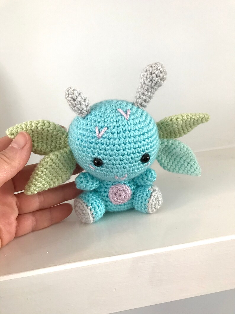 May include: A crocheted blue dragon with green wings and gray horns. The dragon has a pink spot on its belly and black eyes.