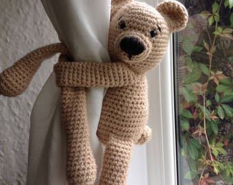 May include: A crocheted teddy bear curtain tieback in a beige color. The bear is holding onto a white curtain with its arms and legs.