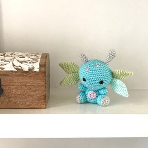 May include: A small, crocheted blue and green creature with large ears and a pink dot on its belly. The creature is sitting on a white shelf.
