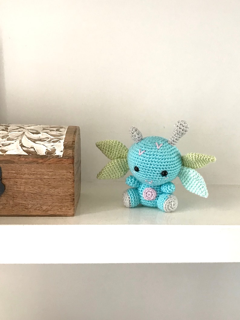 May include: A small, crocheted blue and green creature with large ears and a pink dot on its belly. The creature is sitting on a white shelf.