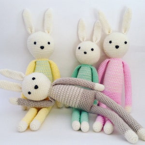 May include: Four crocheted bunny rabbits in different colors. The bunnies are sitting on a white surface. The bunnies are all wearing different colored clothes.