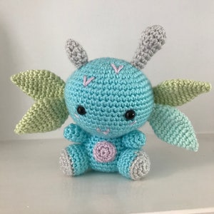 May include: A crocheted blue and green plush toy with gray ears and a pink spot on its belly. The toy has black eyes and a stitched smile.
