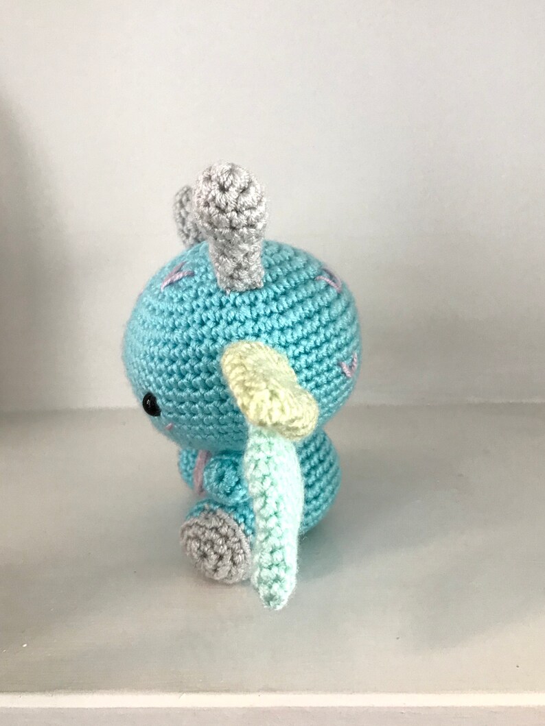 May include: A light blue crocheted stuffed animal with gray and yellow accents. The animal has two horns and a tail.