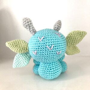 May include: A crocheted blue dragon with green wings and gray horns. The dragon has pink embroidered details on its face.