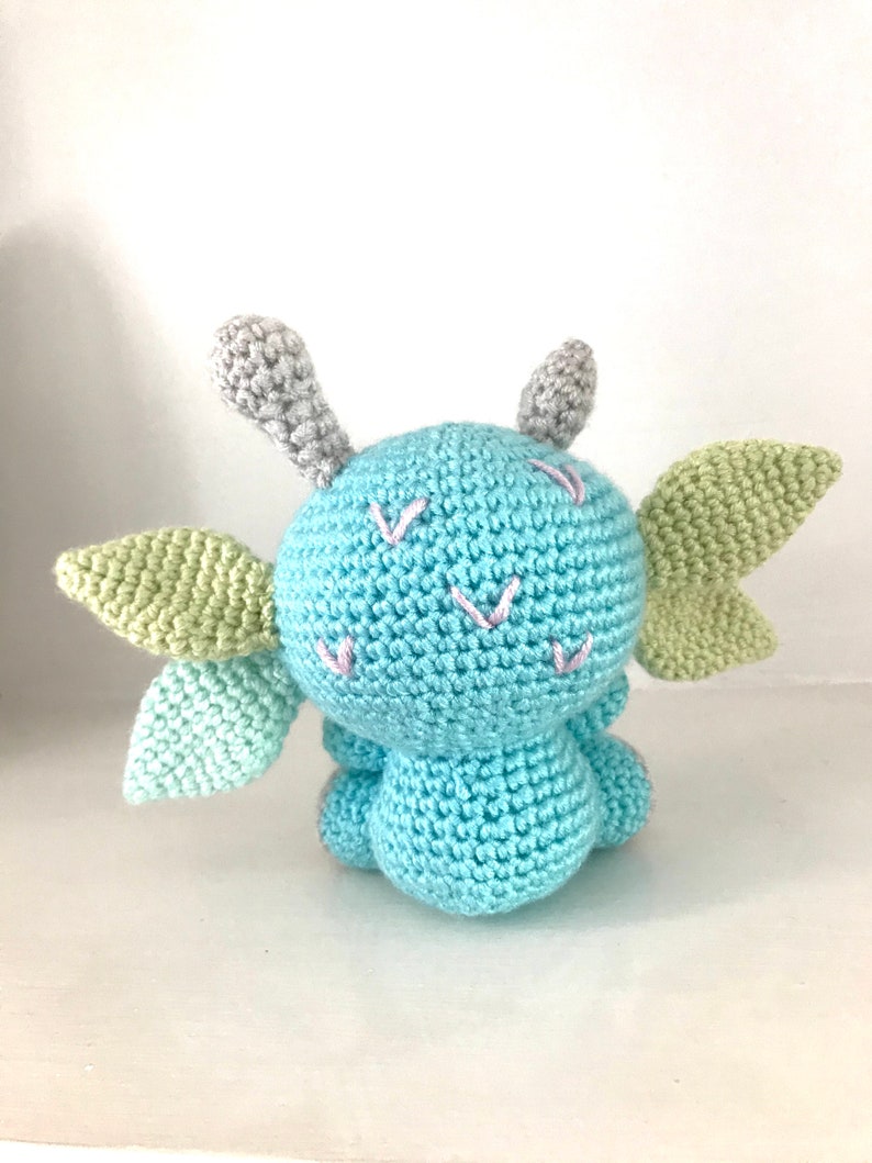 May include: A crocheted blue dragon with green wings and gray horns. The dragon has pink embroidered details on its face.