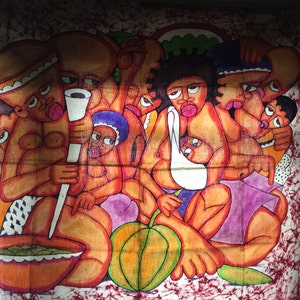 May include: A colorful batik wall hanging depicting a group of people with exaggerated features. The figures are painted in shades of brown, pink, purple, and green, and they are arranged in a circular pattern. The background is a dark red with white swirls.