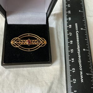 May include: A gold-toned brooch with an oval shape, featuring geometric designs and a central red gemstone, presented in a black jewelry box. The brooch is approximately 2 inches in length.