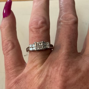 May include: A silver ring with a cluster of square-cut, clear gemstones. The ring is set on a finger with a magenta-colored nail polish. The ring's design features a central row of stones flanked by smaller clusters.