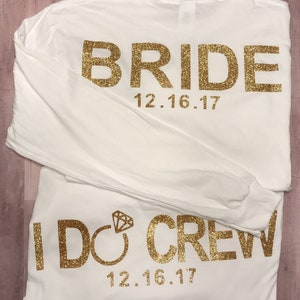 May include: Two white long-sleeved t-shirts with gold glitter lettering. The top shirt says "BRIDE" with the date "12.16.17" underneath. The bottom shirt says "I DO CREW" with the date "12.16.17" underneath.