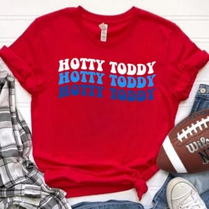 Peut inclure: Un t-shirt rouge avec le texte "HOTTY TODDY" en blanc et bleu. Le t-shirt est associé à un jean bleu, un ballon de football, une chemise à carreaux et des baskets blanches. Le ballon de football est marron avec le texte "Wilson NFL".