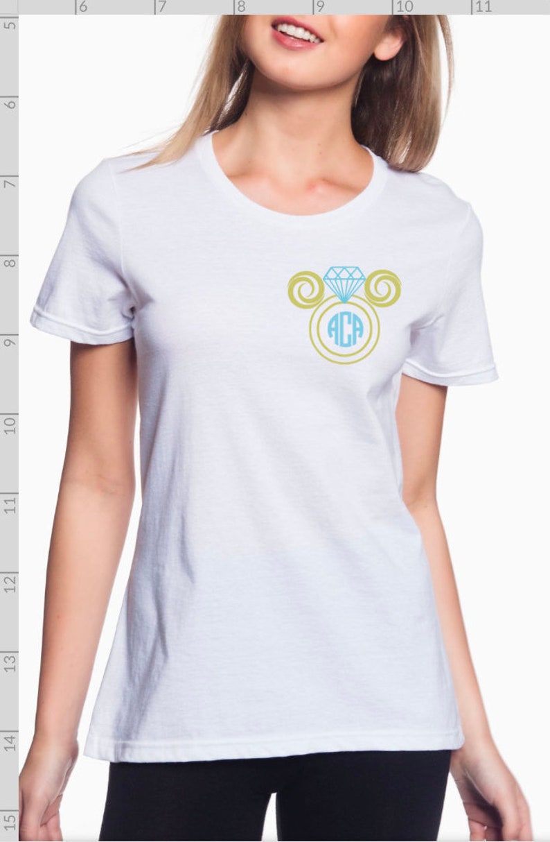 Puede incluir: Camiseta blanca con un anillo de diamante azul claro y remolinos dorados con el monograma "ACA" en el centro.