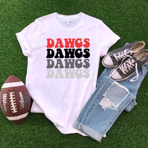 Peut inclure: T-shirt blanc avec le mot "DAWGS" répété quatre fois dans un dégradé de rouge, noir, gris et gris clair. Le t-shirt est posé sur une surface verte avec un ballon de football et une paire de baskets grises.