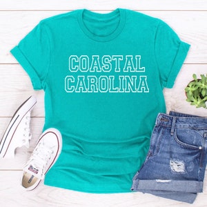May include: A turquoise blue t-shirt with white text that reads "COASTAL CAROLINA". The shirt is laid flat on a white wooden surface with a pair of white sneakers, a pair of blue denim shorts, and a small potted plant.