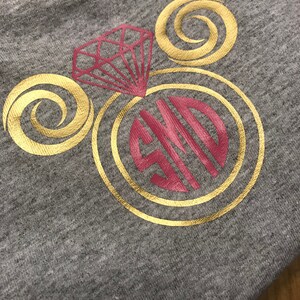 Puede incluir: Una sudadera gris con un dise&ntilde;o dorado y rojo. El dise&ntilde;o presenta un anillo de diamantes con una banda dorada y un diamante rojo. El anillo est&aacute; rodeado por dos remolinos dorados. Las iniciales "SJD" est&aacute;n en el centro del anillo en rojo.
