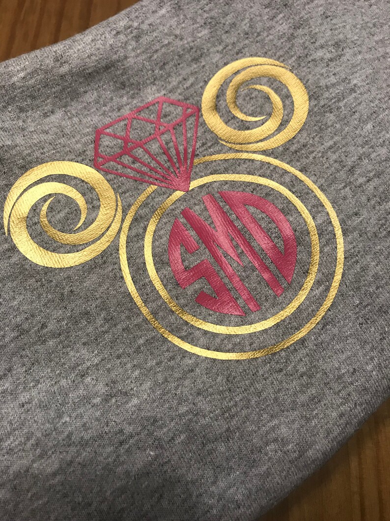 Puede incluir: Una sudadera gris con un dise&ntilde;o dorado y rojo. El dise&ntilde;o presenta un anillo de diamantes con una banda dorada y un diamante rojo. El anillo est&aacute; rodeado por dos remolinos dorados. Las iniciales "SJD" est&aacute;n en el centro del anillo en rojo.