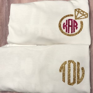 May include: Two white t-shirts with gold glitter monograms. The top shirt has a gold glitter ring with a pink diamond and the monogram "KAR". The bottom shirt has the monogram "YOU".