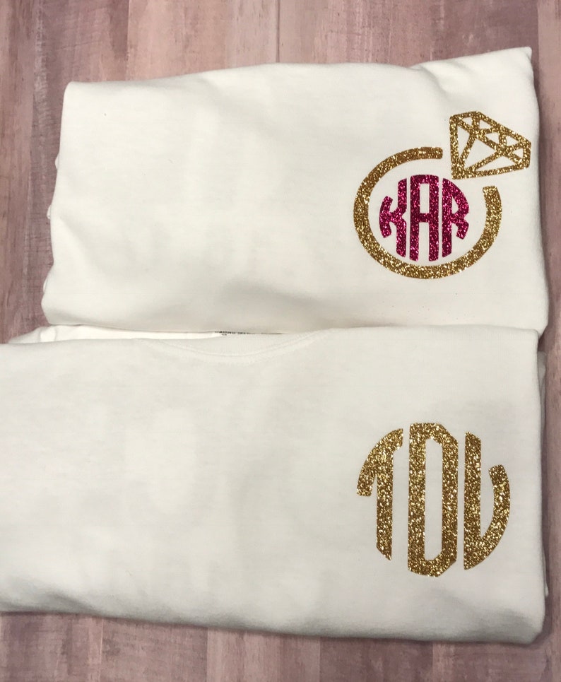 May include: Two white t-shirts with gold glitter monograms. The top shirt has a gold glitter ring with a pink diamond and the monogram "KAR". The bottom shirt has the monogram "YOU".