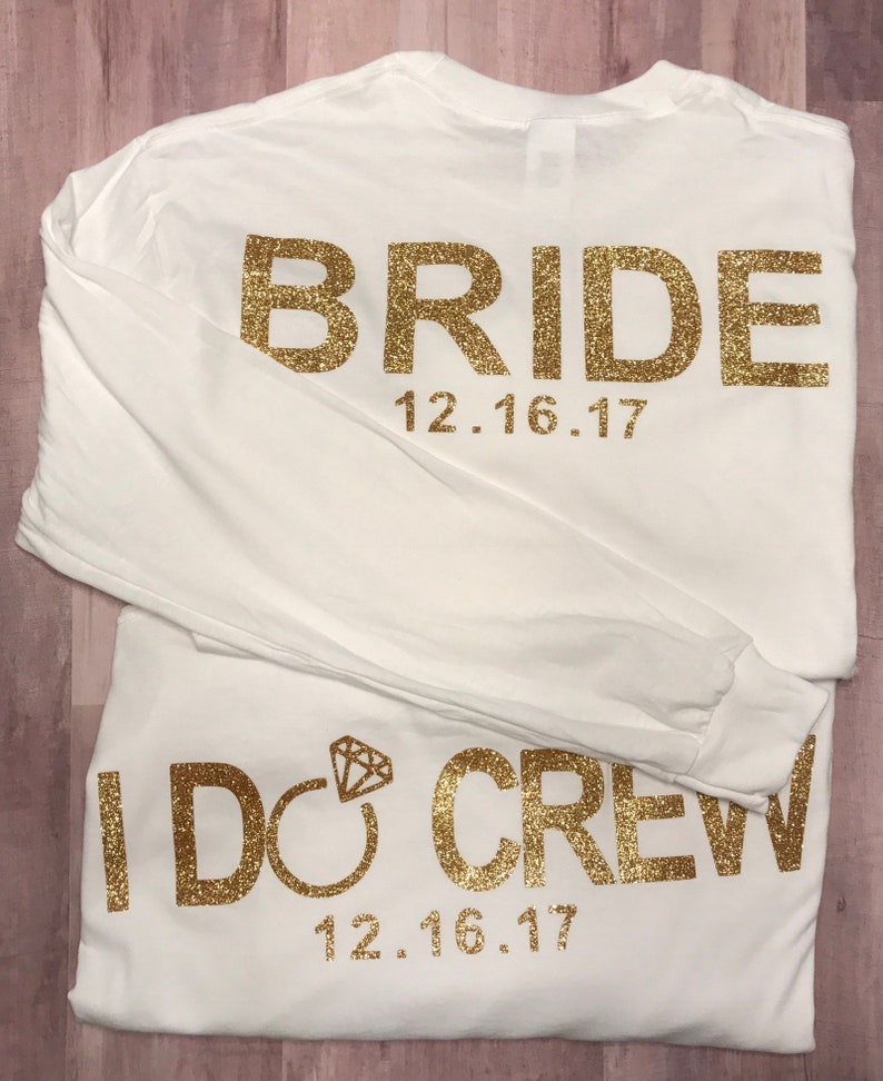 May include: Two white long-sleeved t-shirts with gold glitter lettering. The top shirt says "BRIDE" with the date "12.16.17" underneath. The bottom shirt says "I DO CREW" with the date "12.16.17" underneath.