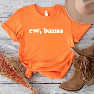 Op de afbeelding: Fel oranje t-shirt met de witte tekst "ew, bama". De afbeelding toont een bruine hoed en bruine enkellaarsjes met franjes. Een bosje gedroogde bloemen maakt de compositie af.