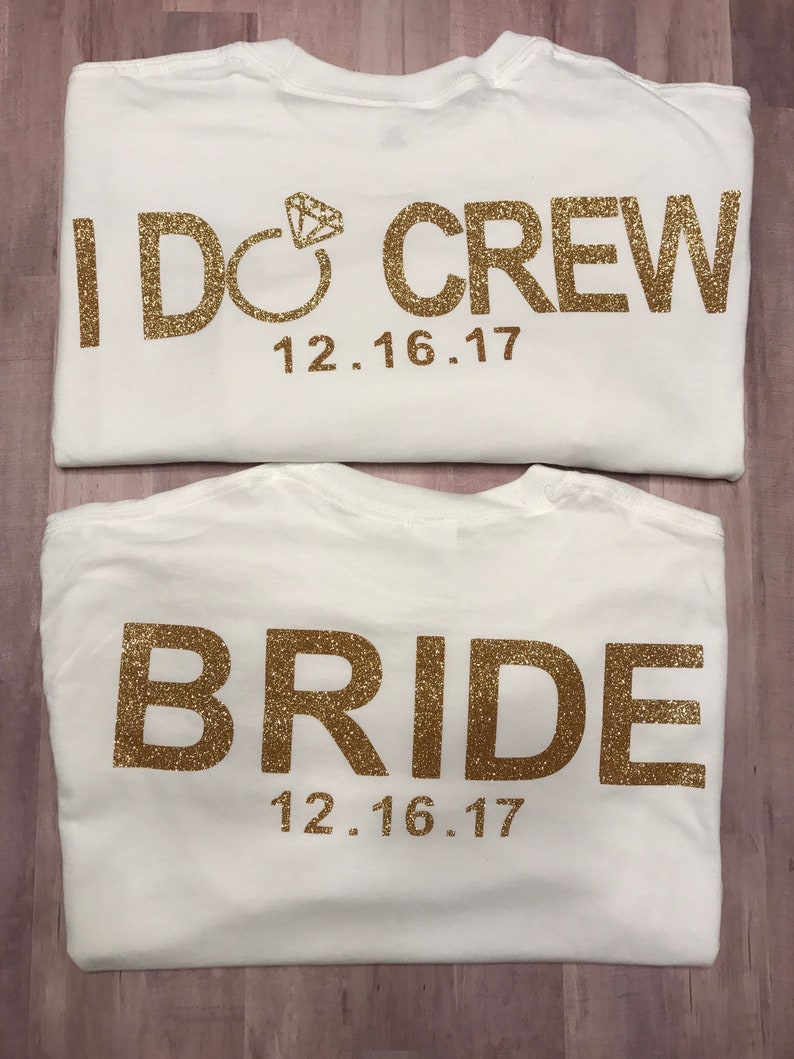 May include: Two white t-shirts with gold glitter lettering. The top shirt says "I Do Crew" with a diamond ring symbol and the date "12.16.17". The bottom shirt says "Bride" with the date "12.16.17".