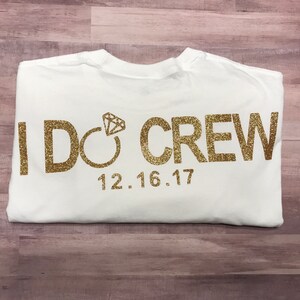 May include: White t-shirt with gold glitter lettering that says "I DO CREW" with a diamond ring graphic and the date "12.16.17".