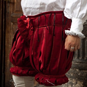 May include: Red velvet breeches with black ribbon ties and a ruffled trim. The breeches are gathered at the waist and have a pleated design.