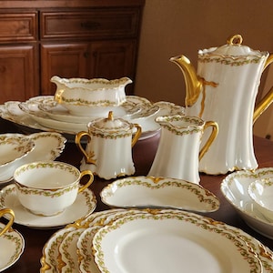 May include: A collection of white porcelain tableware with gold trim, including plates, bowls, cups, saucers, a creamer, sugar bowl, and a coffee pot. The set is arranged on a dark wooden table.