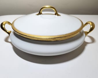 May include: White porcelain serving dish with a lid and gold trim. The dish has two gold handles on the sides and one on the lid. The gold trim features a delicate pattern. A classic piece for serving food.