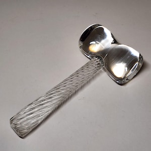 May include: A clear glass hammer with a twisted handle and a double-headed striking surface. The glass is transparent, reflecting light and creating highlights. The handle has a textured, spiral design.