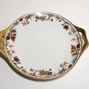 May include: A white porcelain serving plate with gold trim and a floral design in shades of brown, orange, and green. The plate has two gold handles.