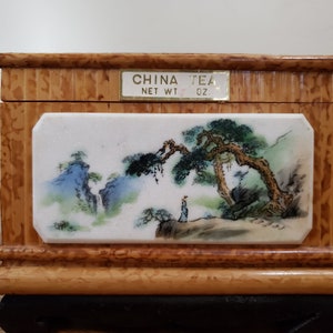 May include: A wooden tea box with a painted porcelain panel depicting a Chinese landscape with a pine tree, mountains, and a person. The box has a gold label that reads "CHINA TEA NET WT. OZ."