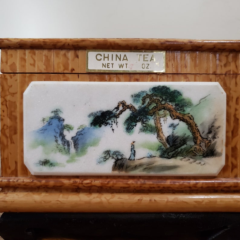 May include: A wooden tea box with a painted porcelain panel depicting a Chinese landscape with a pine tree, mountains, and a person. The box has a gold label that reads "CHINA TEA NET WT. OZ."